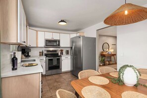 A modern kitchen with white cabinets, a stainless steel refrigerator, a microwave, and an oven. There is a dining area with a wooden table and woven chairs. - Hot Tub, Sauna & Game Room Mountain-View Home (Westminster)