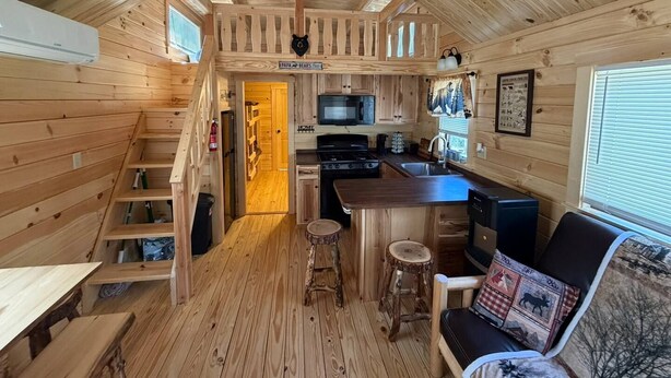 A wooden cabin interior with a kitchen, dining area, and a staircase leading to a loft.