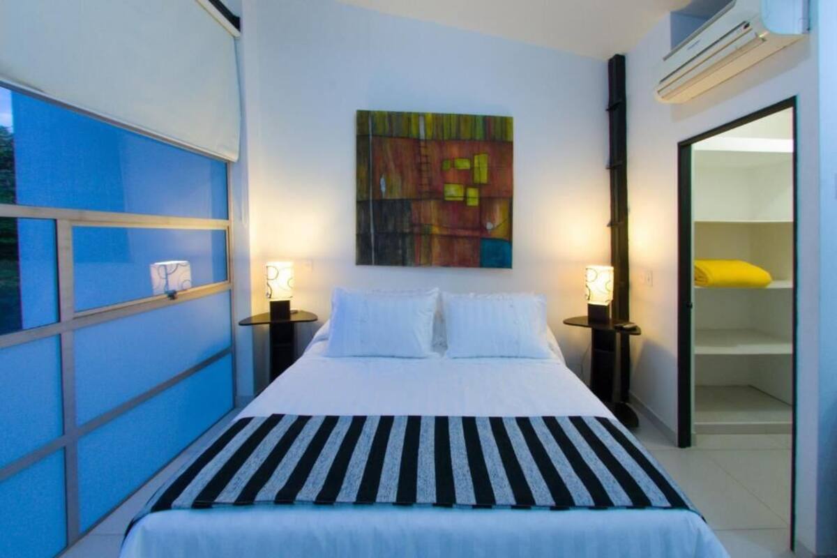 A hotel room with a bed, two bedside tables with lamps, a colorful abstract painting on the wall, and a sliding door leading to a bathroom.