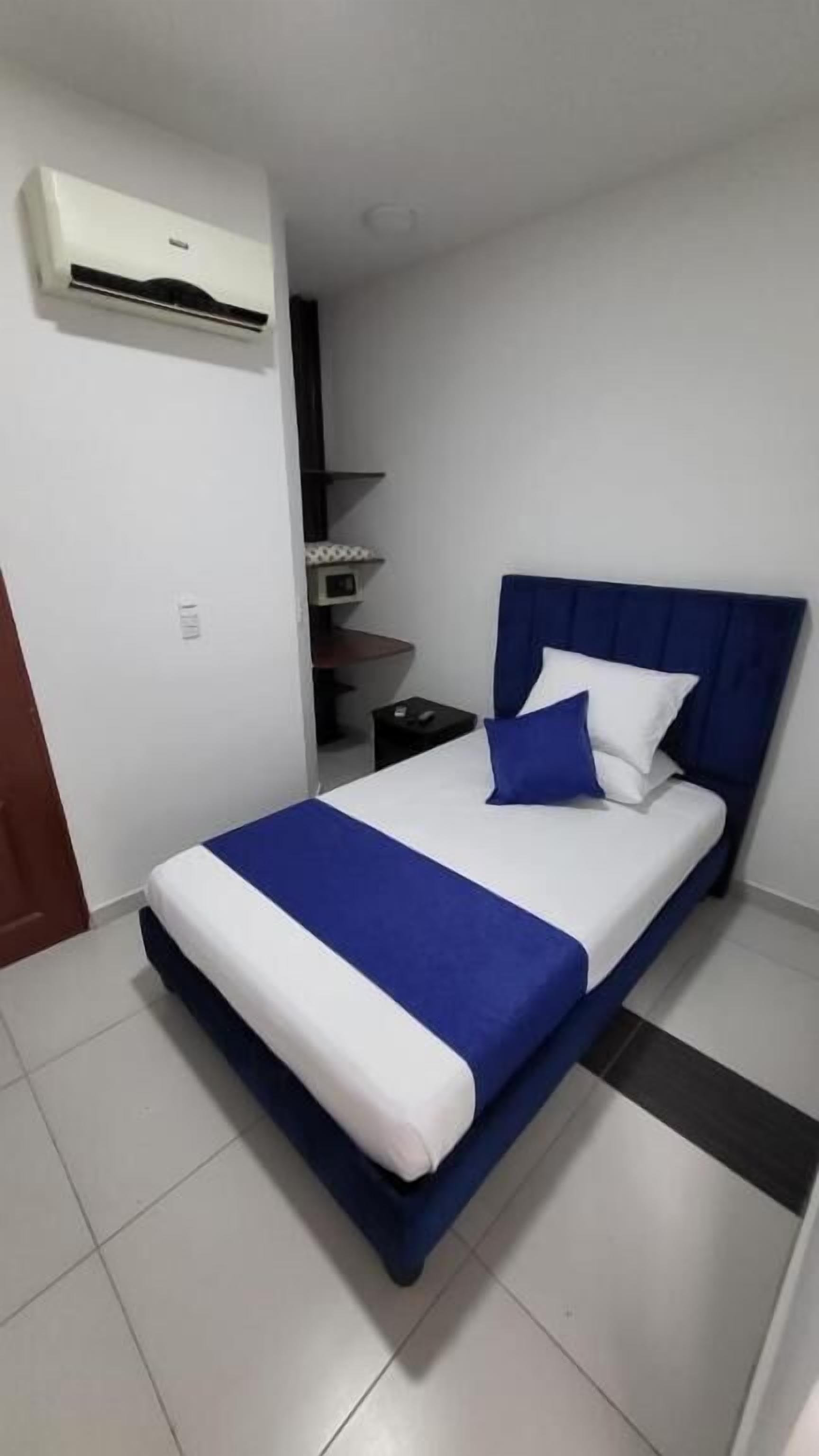 A single bed with a blue headboard and white bedding, a bedside table, a nightstand, and an air conditioning unit mounted on the wall.