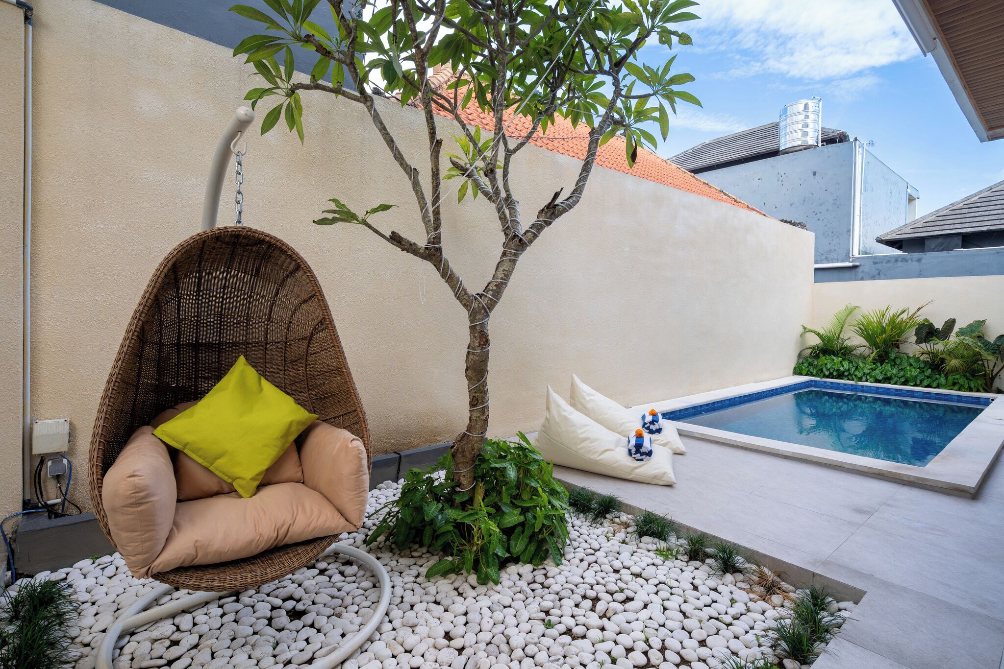 A hanging chair with cushions, a small tree, a white wall, and a swimming pool.