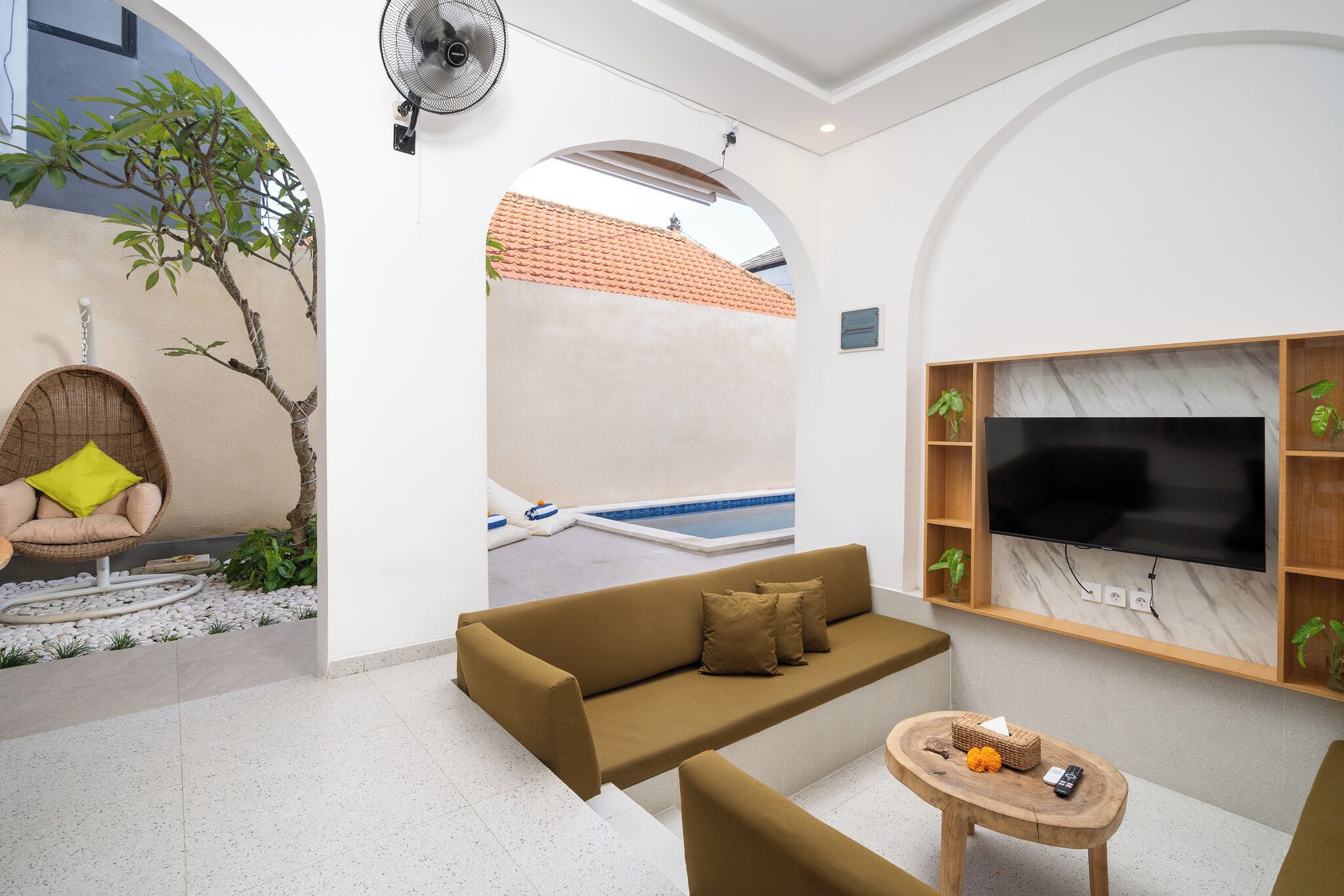 Ein modernes Wohnzimmer mit einer Couch, einem kleinen runden Tisch, einem an der Wand montierten Fernseher und Blick auf einen Swimmingpool durch einen Bogen.