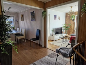 A modern living room with a dining area, wooden flooring, a TV, and various plants.