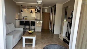 Ein moderner Wohnbereich mit Sofa, Esstisch, Stühlen und einer Küche im Hintergrund. Ein Fernseher befindet sich rechts.