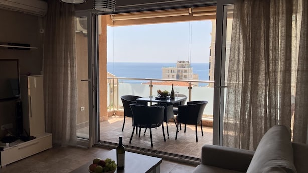 Ein Wohnzimmer mit einer Couch, einem Couchtisch mit Obst, einem Essbereich mit Stühlen und einem Tisch sowie ein Balkon mit Meerblick.