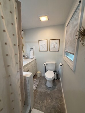 Un baño con inodoro, lavamanos y cortina de ducha. Hay dos cuadros enmarcados en la pared y una ventana con persianas.
