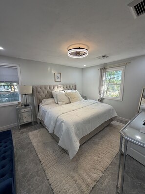 Ein Schlafzimmer mit einem großen Bett, Nachttischen, einer Chandelüster, einem Fenster mit Vorhängen und Blick nach draußen.