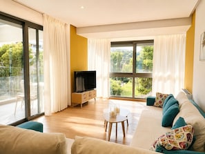 A modern living room with a sofa, a TV on a wooden cabinet, a small coffee table, and large windows with curtains.