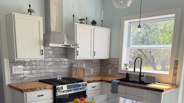 A modern kitchen with white cabinets, a marble island, and a large window. - The Still Waters Cottage (Evart)