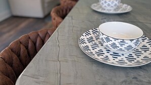 A dining table with patterned plates and a cup, a brown woven bench, and a light-colored sofa in a room with a window. - The Still Waters Cottage (Evart)