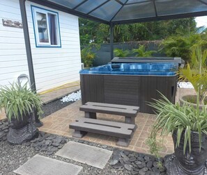 A hot tub in a covered outdoor area with a wooden bench, surrounded by potted plants and a stone pathway. - BUNGALOW DE SAINT JACQUES (Saint-François)