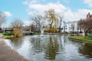 Ein stiller Teich mit einer Fontäne, umgeben von kahl stehenden Bäumen und einem weißen Gebäude.