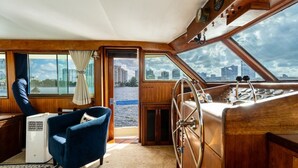 A boat interior with a steering wheel, a blue chair, a small table, and a view of the city through the windows. - Stay Aboard a Vintage Hatteras 53' Motor Yacht in Aventura-A Unique Experience! (Aventura)