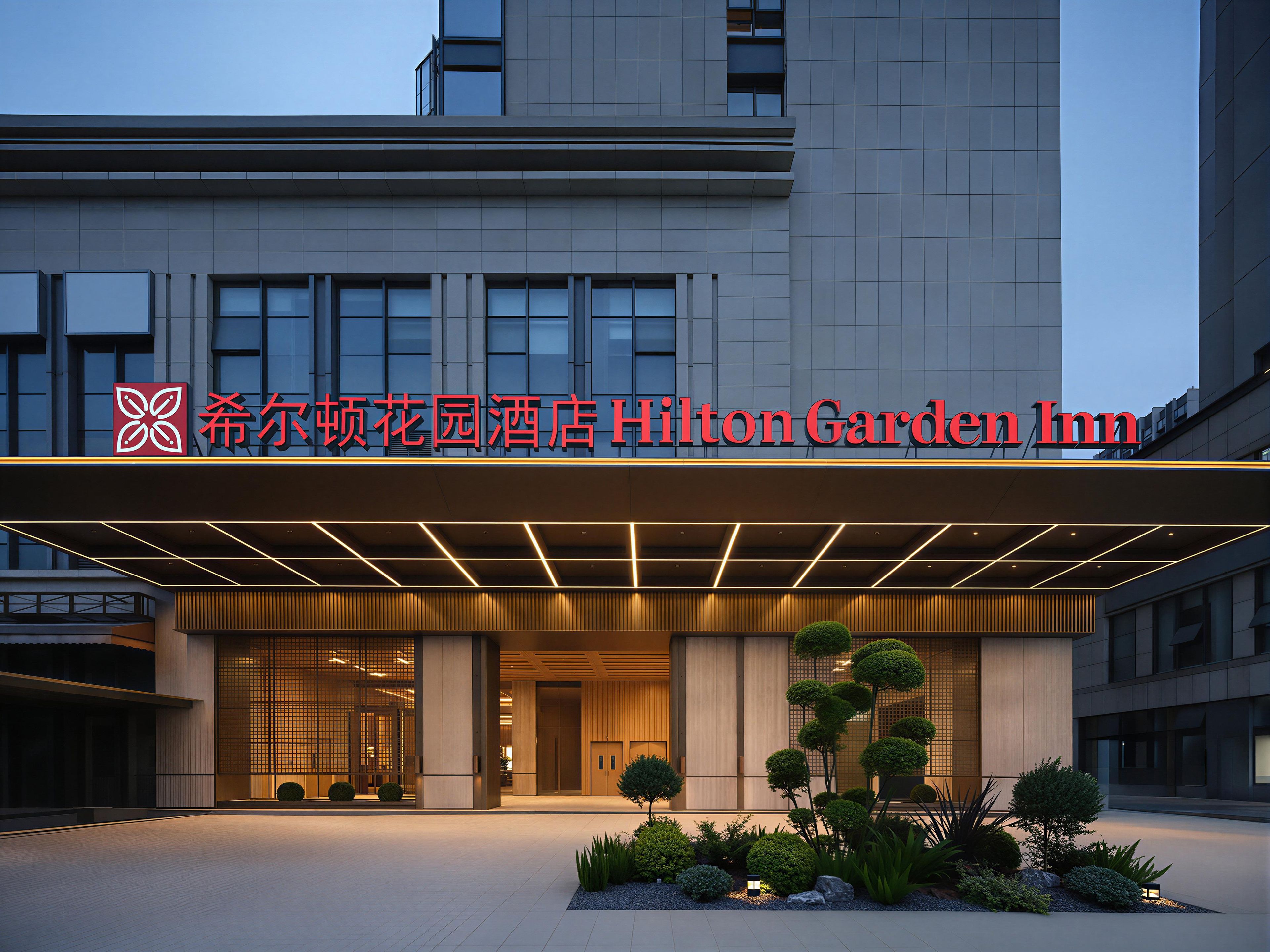 A hotel entrance with a modern facade, a prominent sign, and a landscaped garden area.