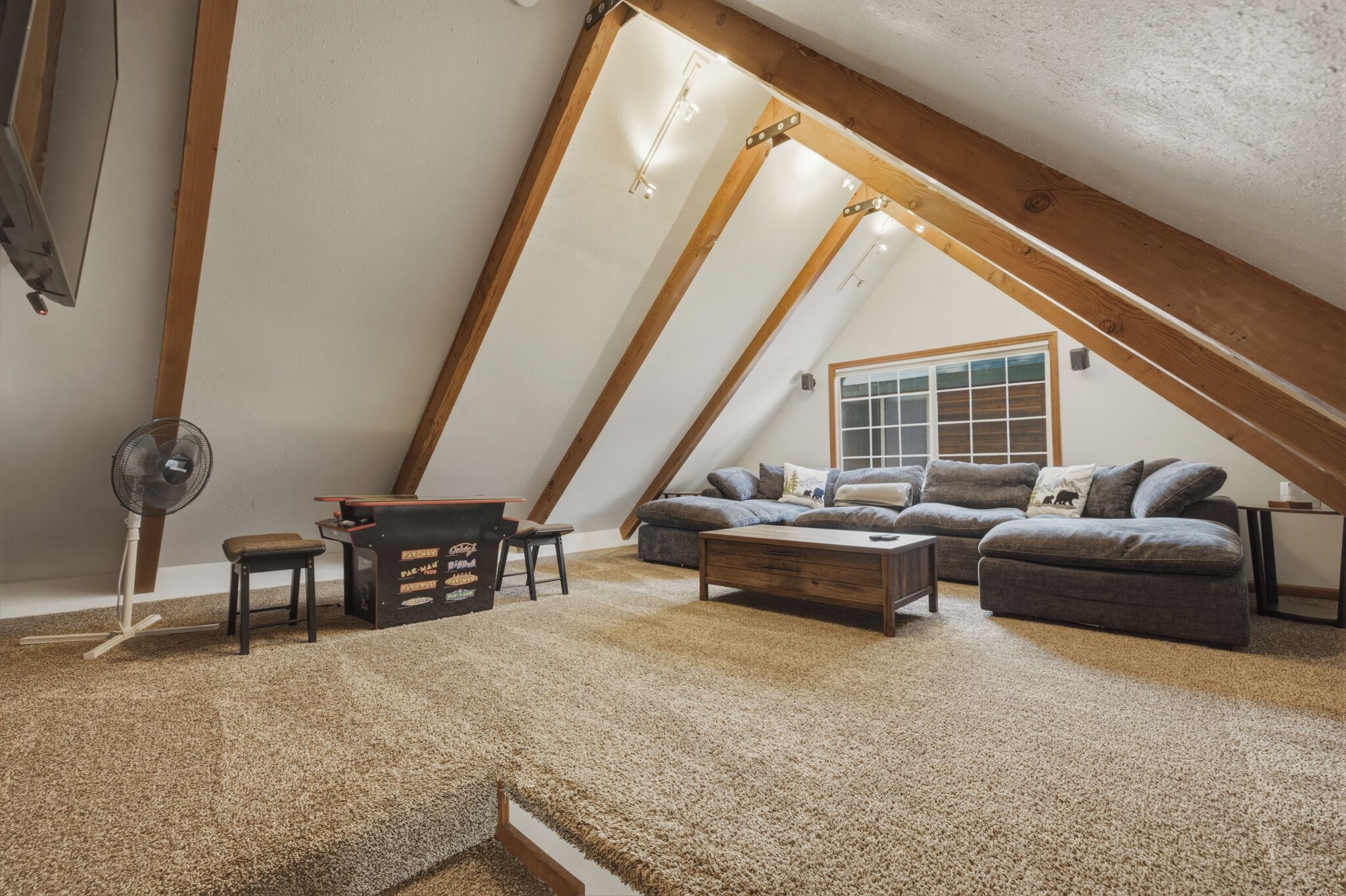 A cozy attic room with a sloped ceiling, a large window, a sofa set, a coffee table, and a fan.