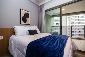 A hotel room with a bed, a framed artwork on the wall, and a view of a balcony with buildings. - Modern studio in the heart of Vila Madalena. (São Paulo)