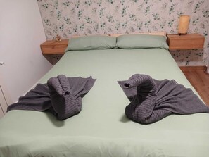 Un lit recouvert d’une couverture verte, deux serviettes pliées, une table de chevet en bois avec une lampe, et un papier peint à motifs floraux en arrière-plan.