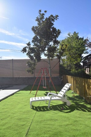 Een achtertuin met kunstgras, een schommelset, een witte loungestoel en een houten omheining.