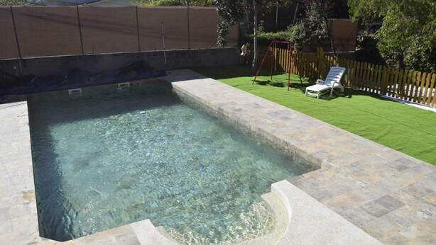 A rectangular swimming pool with clear water, surrounded by stone tiles and a grassy area with a wooden fence and a swing set.