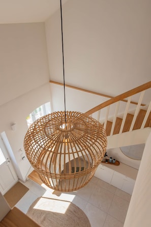 Ein modernes Interieur mit einer hölzernen, gewebten Leuchte, die von der Decke hängt, einer Treppe mit Holzgeländern und einem Blick ins Freie durch ein Fenster.