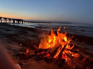 A beach scene with a fire, a pier in the background, and a clear sky. - Die Alte Meierei (Steinbergkirche)