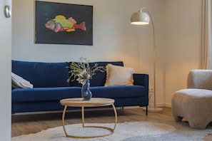 Ein Wohnzimmer mit einem blauen Sofa, einem runden Couchtisch, einer Stehlampe und einem Bild von einem Fisch an der Wand.