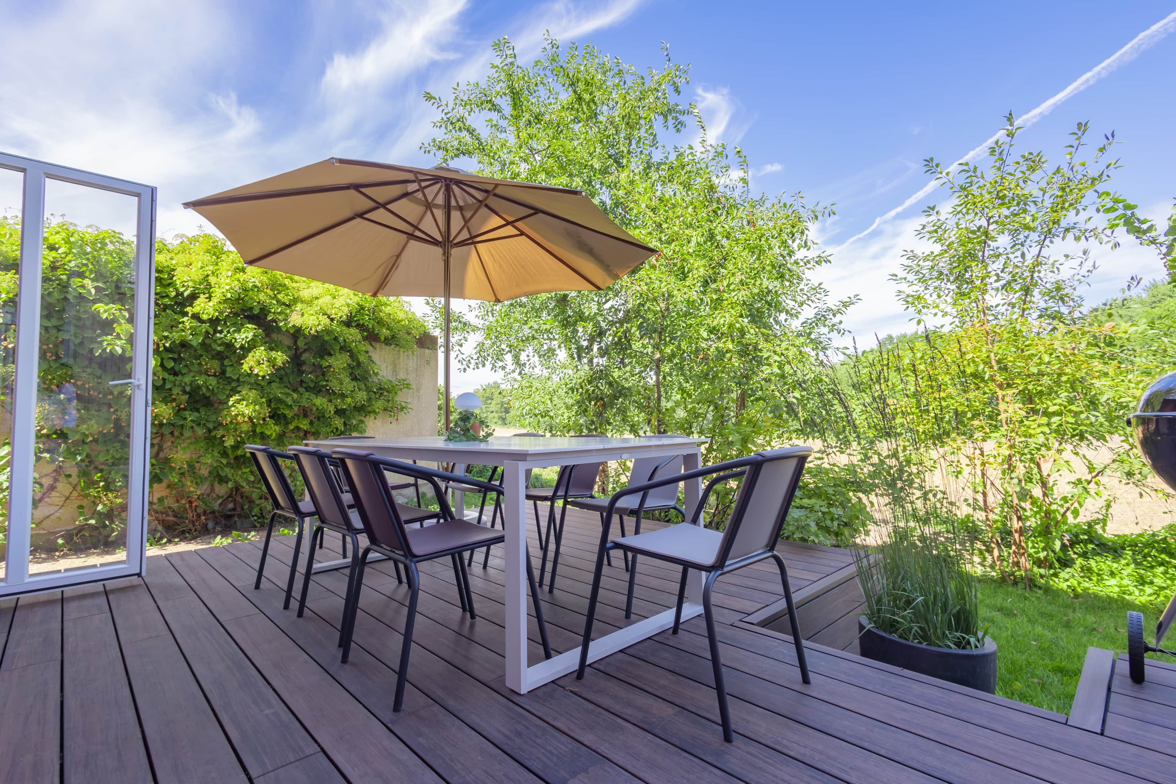 An outdoor patio with a table and chairs, a large umbrella, and a view of greenery.