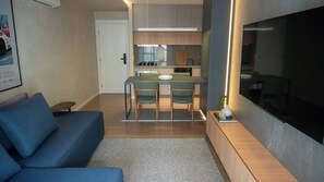 A modern living room with a blue sofa, a dining area with chairs, a kitchen with wooden cabinets, and a large TV on the wall.