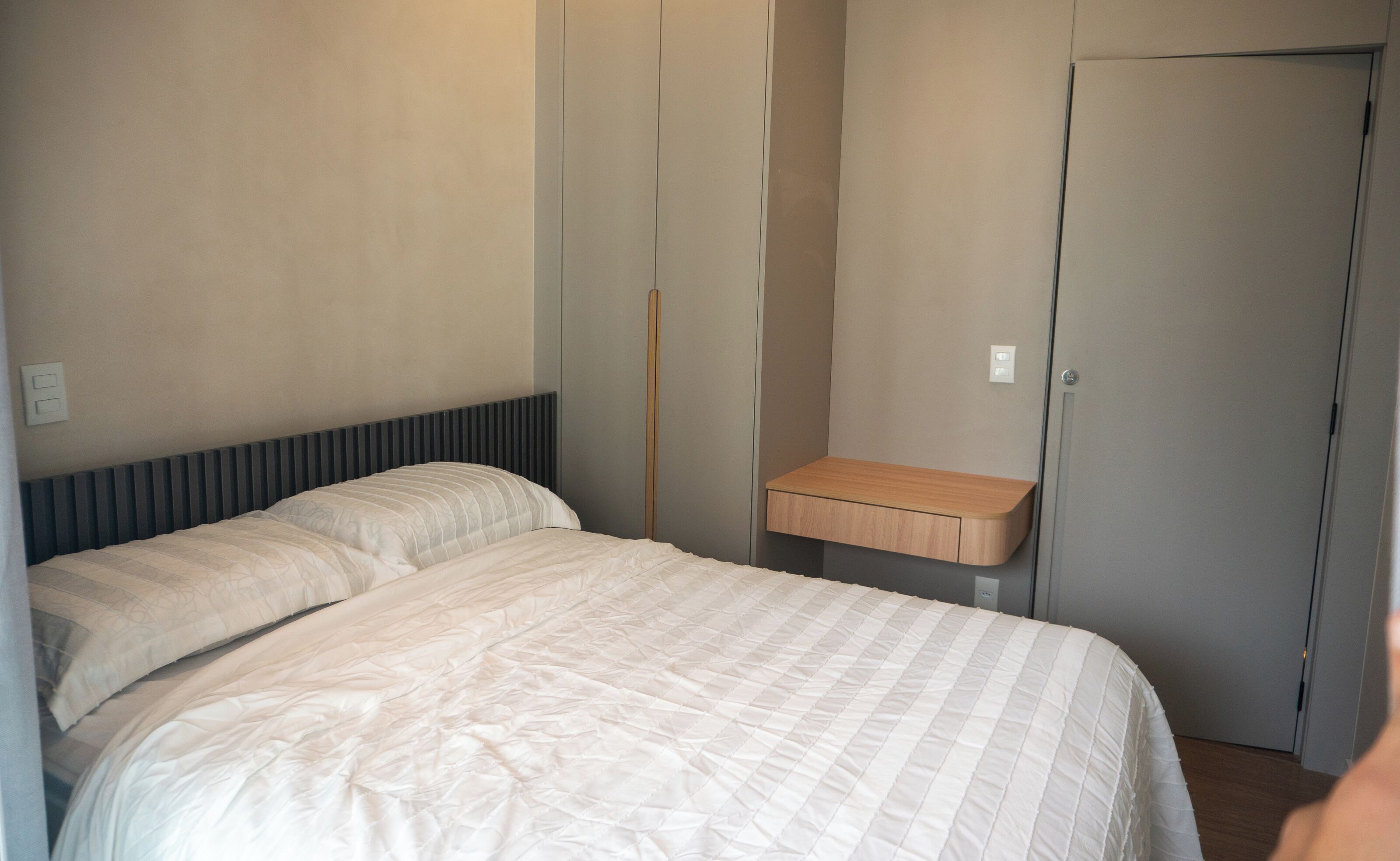 A bedroom with a bed, a bedside table, a wardrobe, and a wall-mounted light switch.