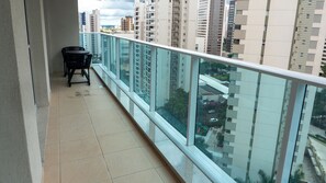 Un balcon doté d’une balustrade en verre, d’une petite table et de chaises, offrant une vue sur un paysage urbain avec de nombreux immeubles de grande hauteur.