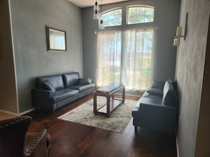 Sala de estar con un sofá de color oscuro, una mesa de centro y una alfombra. Se ven dos ventanas con cortinas y una lámpara de araña.