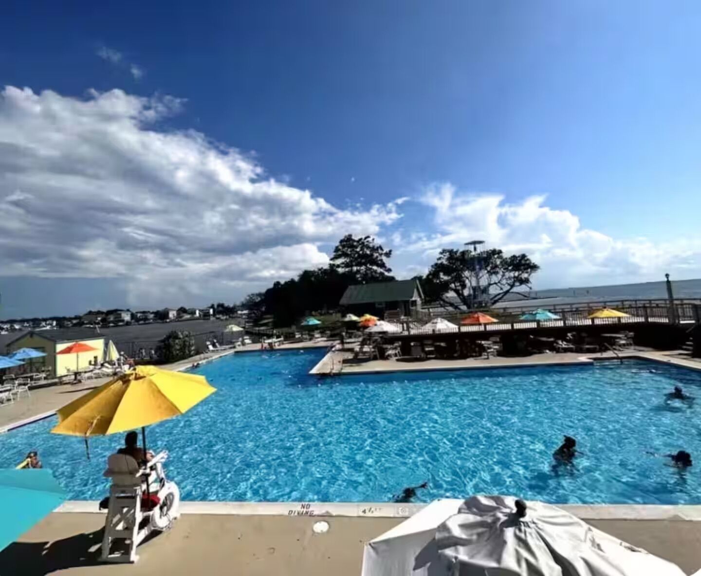 Une piscine avec des personnes dans l’eau, un poste de surveillance pour les sauveteurs et des parasols.