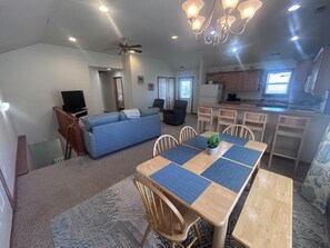 A living room with a sofa, TV, and ceiling fan; a dining area with a table and chairs; and a kitchen with cabinets and a refrigerator. - Lily's Pad (Kill Devil Hills)