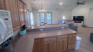 A kitchen with wooden cabinets, a countertop, a ceiling fan, a television, and a dining area with a chandelier. - Lily's Pad (Kill Devil Hills)