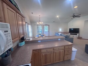 A kitchen with wooden cabinets, a countertop, a ceiling fan, a television, and a dining area with a chandelier.