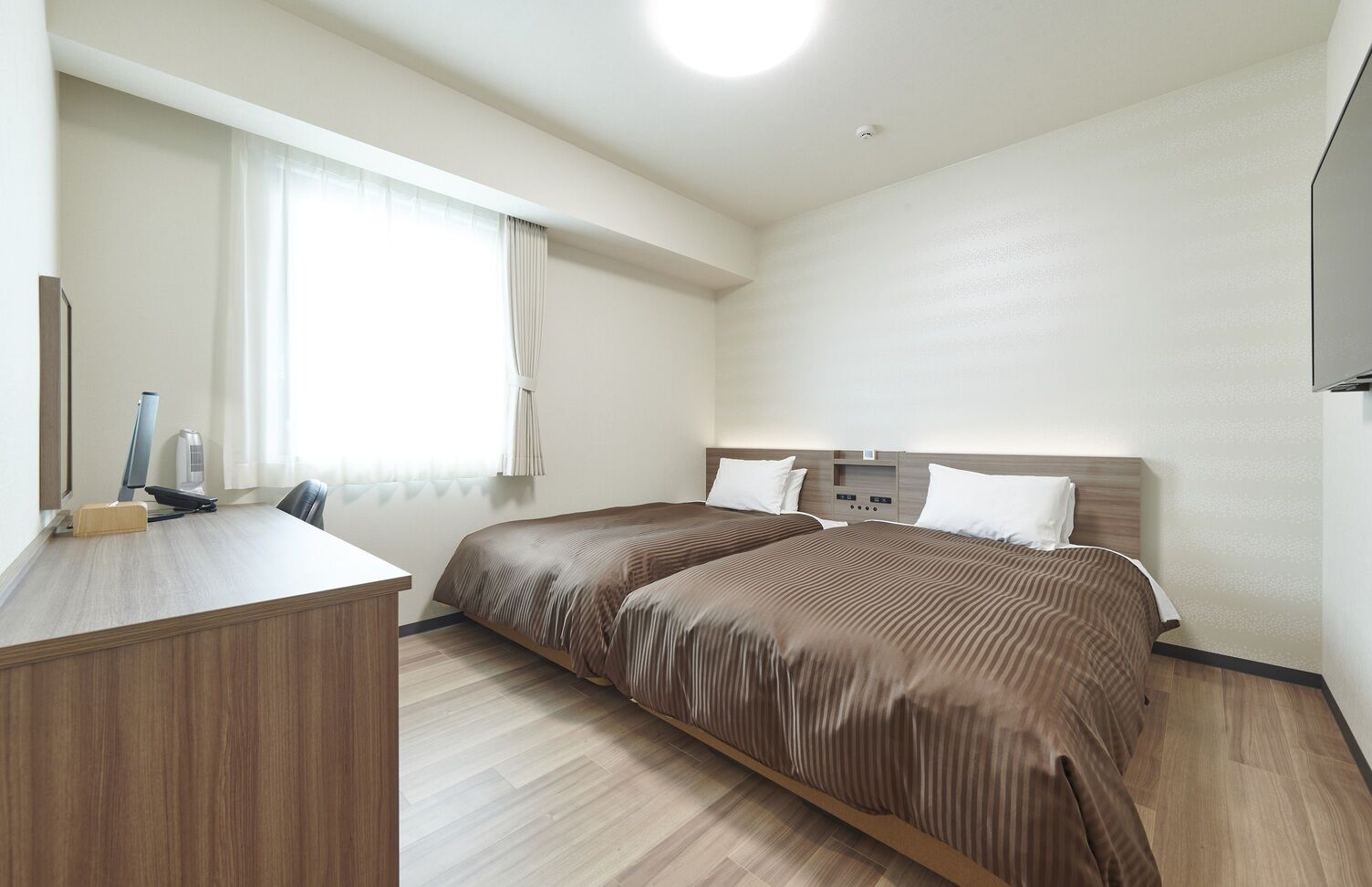 A hotel room with a wooden desk, two beds with brown bedding, a small bedside table, and a window with curtains.