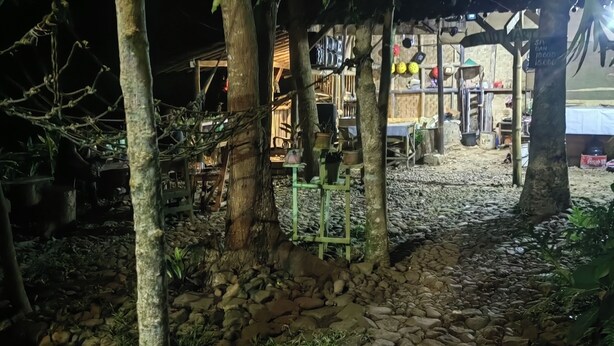 Uma área externa à noite, com árvores, um caminho de pedra e uma estrutura com prateleiras e objetos.