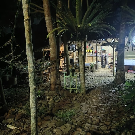 A nighttime outdoor area with trees, a stone pathway, and a structure with shelves and items.. Gazebo Riverside Cibolang By Arsakha