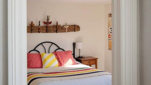 A bedroom with a bed, pillows, a nightstand, a lamp, a wall-mounted shelf with decorative items, and a patterned rug. - Maison Bloomfield - Centrally Located Charming Gem (Prince Edward)