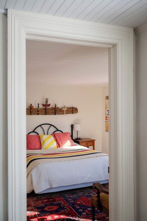 A bedroom with a bed, pillows, a nightstand, a lamp, a wall-mounted shelf with decorative items, and a patterned rug.