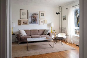 A living room with a sofa, coffee table, and a chair. There are framed pictures on the wall and a window with curtains.