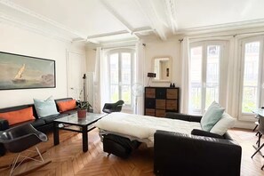Ein geräumiges Zimmer mit einem Bett, einem Sofa, einem Esstisch und einem Stuhl. Ein großes Fenster mit Vorhängen, ein Bild an der Wand und ein Spiegel über dem Bett sind vorhanden.