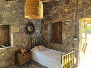 A bedroom with stone walls, a bed with a white bedspread, a wooden nightstand, a wicker lamp, and a mirror with a sunburst design. - Casa Paz by Otium (La Cruz)