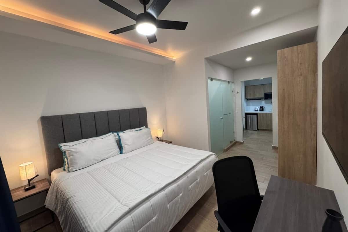 Ein Schlafzimmer mit Bett, Nachttischen, einem Schreibtisch, einem Sessel und einem Deckenventilator, mit Blick zur Küche.