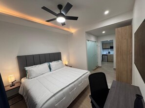 A bedroom with a bed, bedside tables, a desk, a chair, a ceiling fan, and a view into the kitchen.