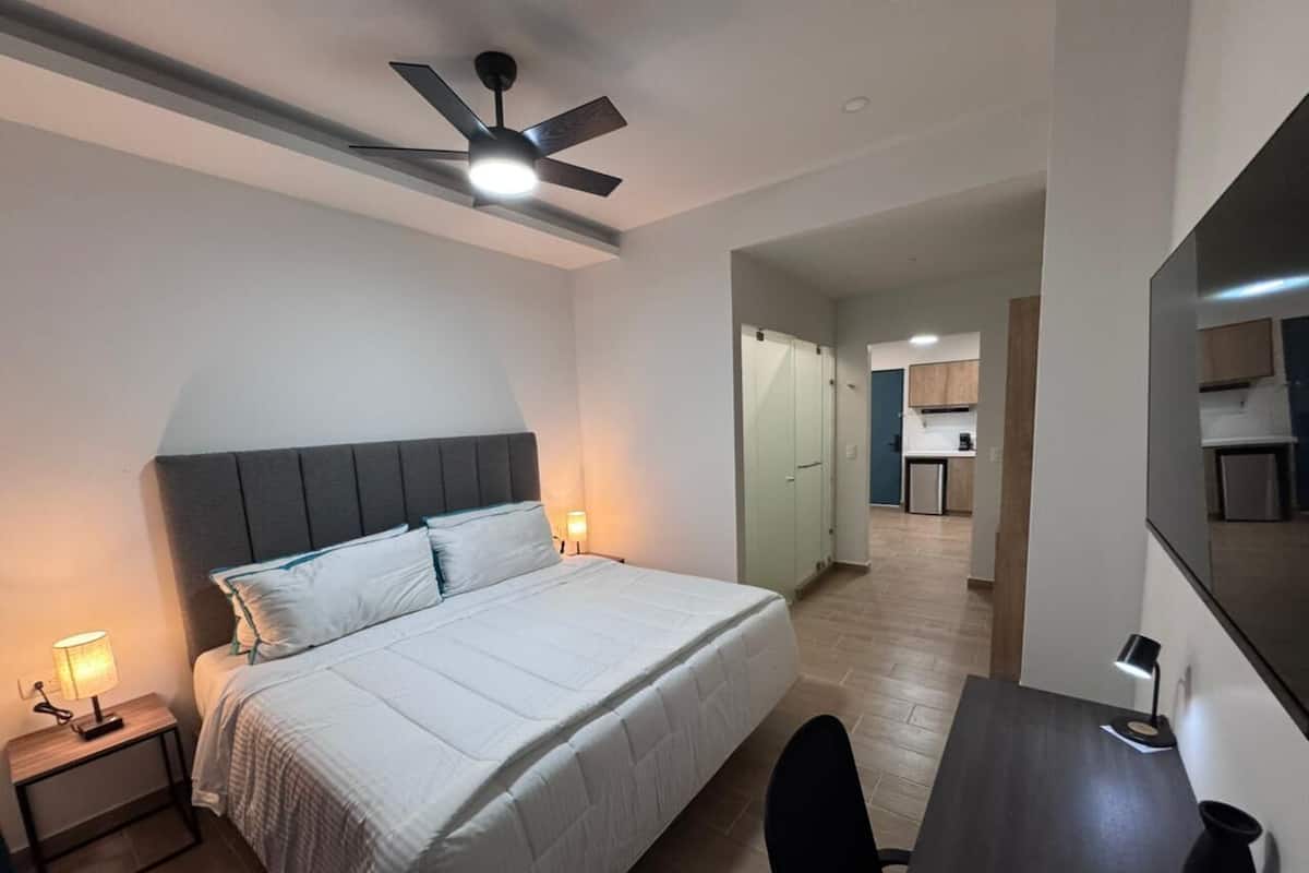 Ein Hotelzimmer mit Bett, Nachttischlampen, Schreibtisch, Stuhl, Deckenventilator und Blick in den Küchenbereich.