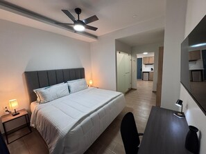 A hotel room with a bed, bedside lamps, a desk, a chair, a ceiling fan, and a view into the kitchen area.