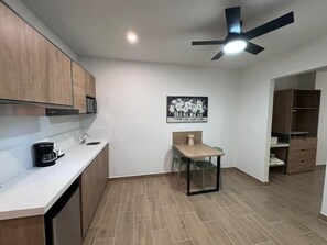 A modern kitchen with wooden cabinets, a coffee maker, a dining table with chairs, a ceiling fan, and a framed picture on the wall.
