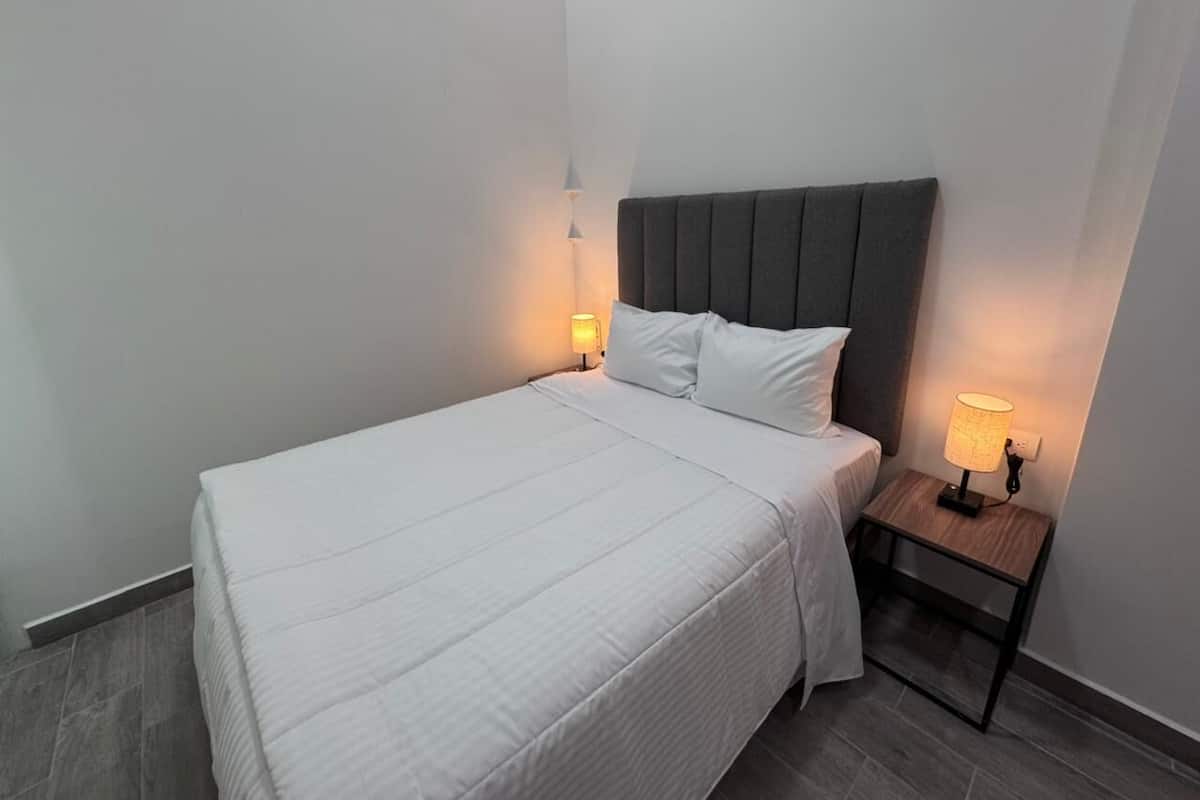 Ein ordentlich gemachtes Bett mit weißer Bettwäsche, einem grauen Kopfteil und zwei Nachttischlampen auf einem Holznachttisch.