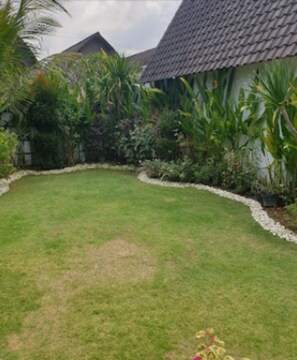 A well-maintained lawn with a stone border, surrounded by various tropical plants and a building with a tiled roof. - Cozzy  White House Villa Bali (Badung)